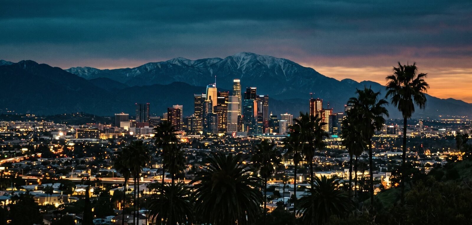 Downtown Los Angeles at dusk behind palm trees, with the San Gabriel Mountains beyond