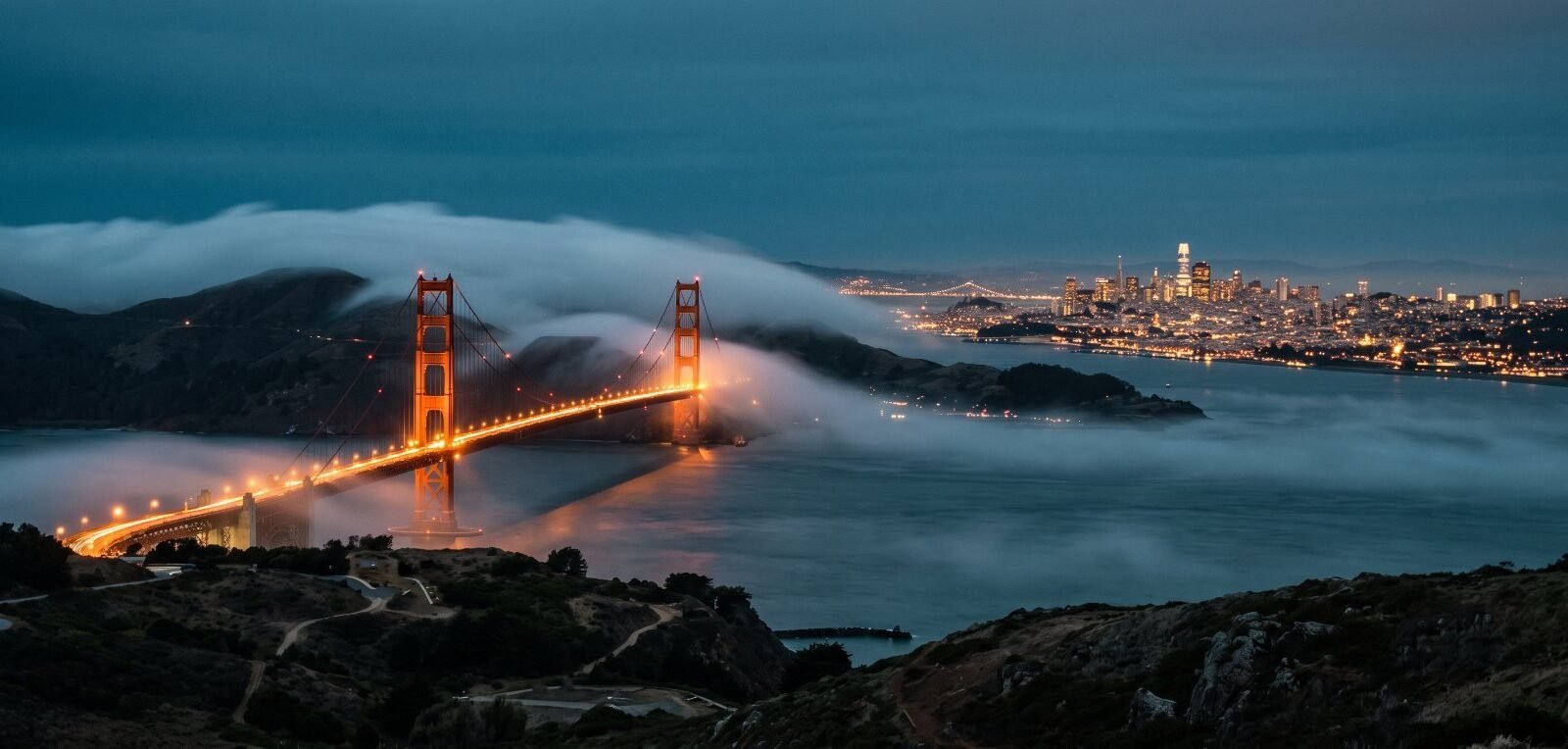Fog rolling over the Golden Gate Bridge at dusk with San Francisco lit behind it